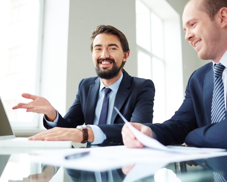 Image of smiling businessman pointing at laptop screen while explaining ideas to his colleague at meeting and looking at camera
